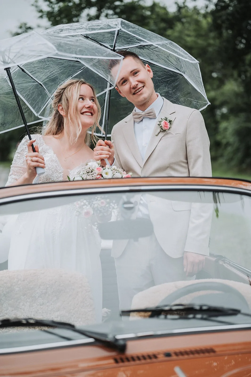 Lina und Johannes lachen unter einem durchsichtigen Regenschirm hinter einem orangefarbenen Oldtimer-Cabrio